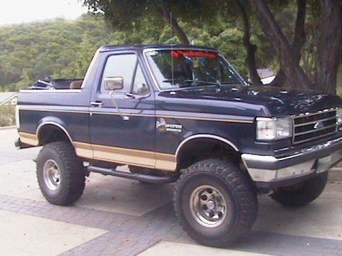 1991 Ford Bronco - Dark blue lifted Ford full-size Bronco with two-tone trim and chrome wheels, side profile on a paved driveway under trees.
