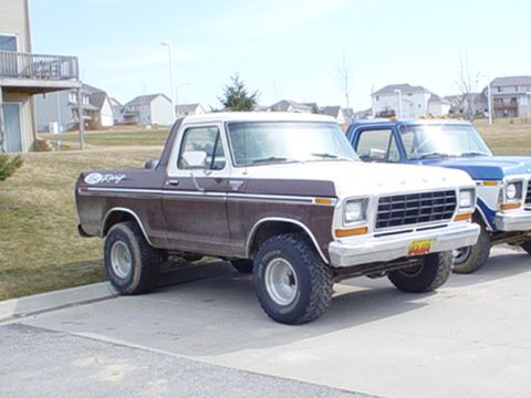 1979 Ford Bronco - A two-tone brown and white 1979 Ford Bronco is parked in front of a blue truck and residential buildings.