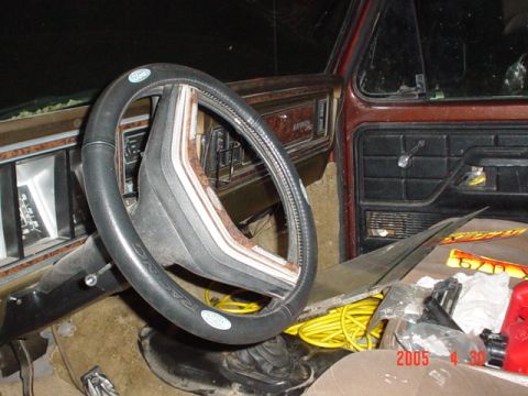 1979 Ford Bronco - The interior of a vehicle shows a steering wheel, dashboard, and door panel with various items on the floor.