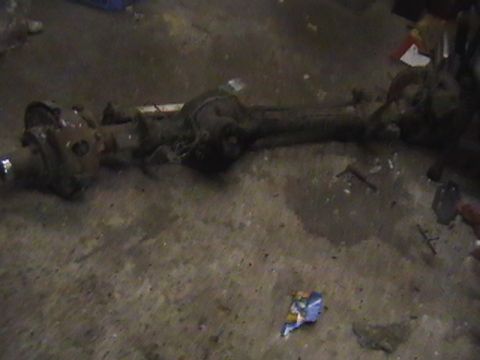 1986 Ford Bronco - A rusty rear axle assembly lies on a dirty concrete floor, with debris scattered around.