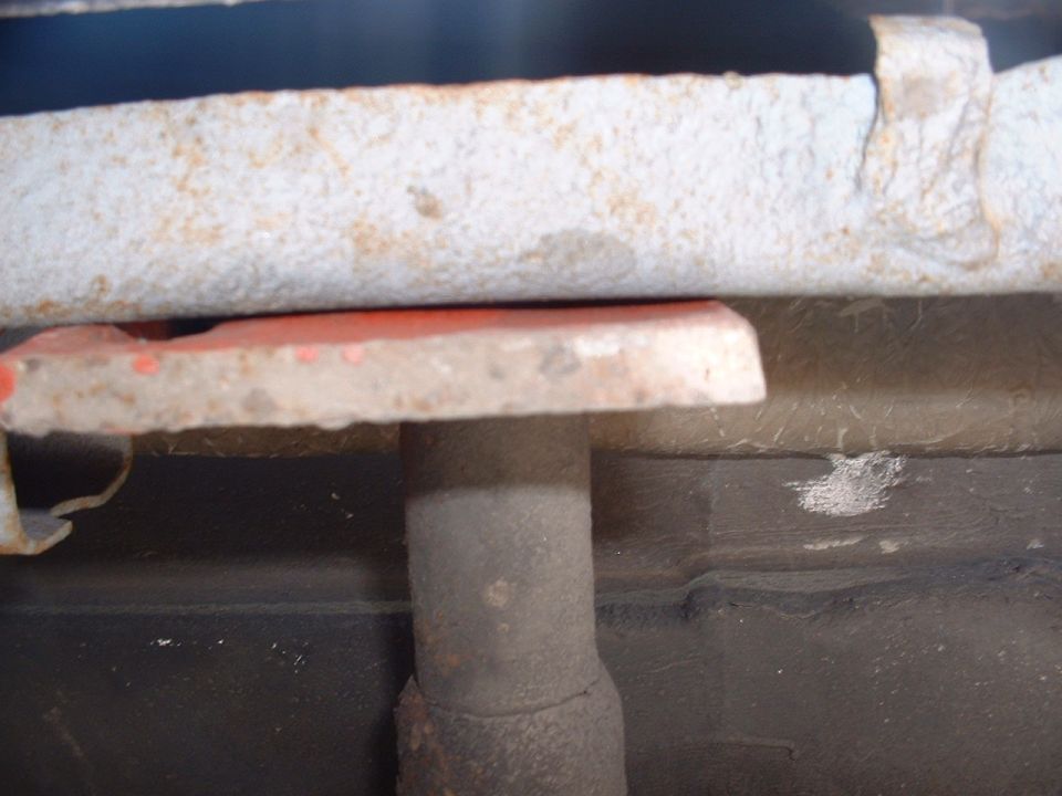 Underside view of a rusty metal component with an exhaust pipe below, showing a red-painted bracket