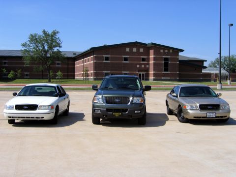 2000 Ford Crown Victoria - Three Ford vehicles are parked in a lot in front of a school building on a sunny day.