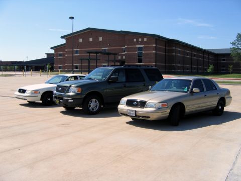 2000 Ford Crown Victoria - Three vehicles, including a gold Ford Crown Victoria, are parked in a lot outside a brick building.