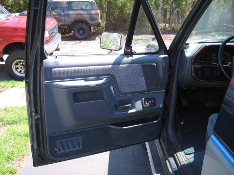 1979 Ford Bronco - View of the interior door panel and dashboard, with a red truck and a camouflage SUV parked nearby.