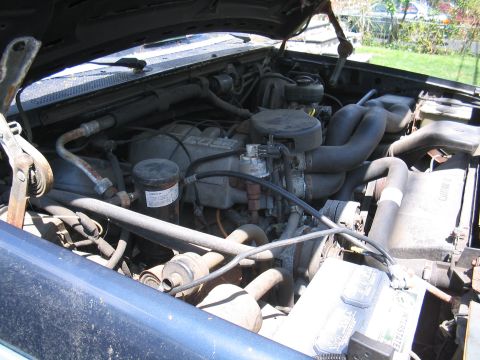 1979 Ford Bronco - The engine bay of a blue vehicle is shown, with various hoses, the battery, and engine parts visible.
