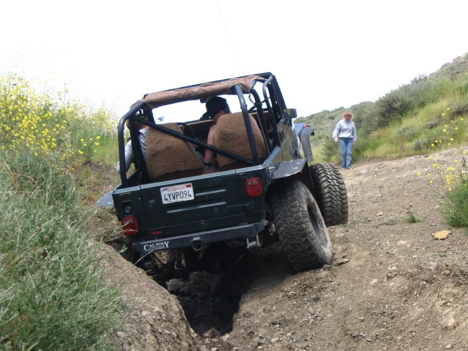 Rear of green soft-top Jeep flexed in a deep dirt rut, Cal Poly bumper sticker visible