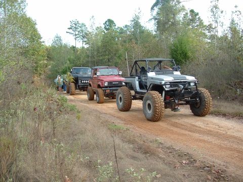 1978 Ford Bronco - Modified off-road vehicles traverse a dirt path surrounded by trees.