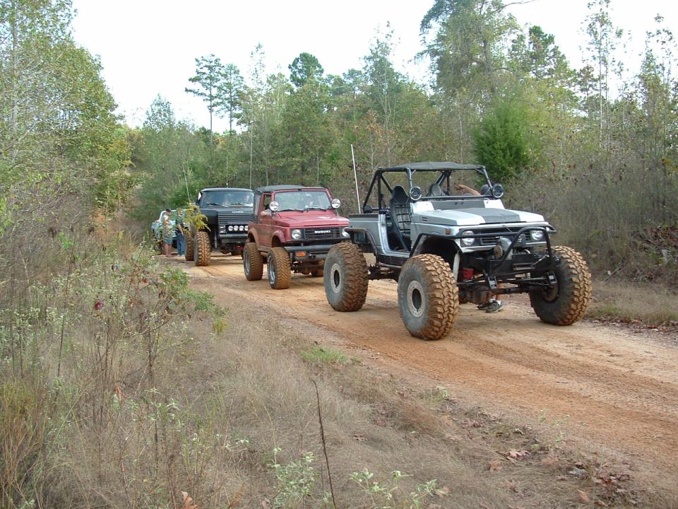 Four modified off-road vehicles driving in a line on a dirt road through a wooded area