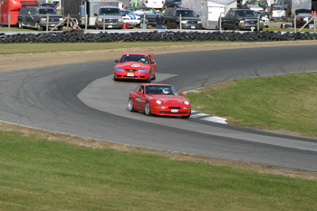 2000 Ford Mustang - Two red sports cars compete on a race track, with one car leading the other around a curve.
