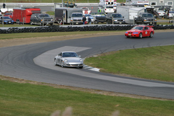 2000 Ford Mustang - A silver Porsche 911 navigates a turn on a race track, with other vehicles visible in the background.