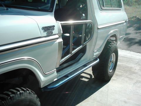 1978 Ford Bronco - Close side view of a white square-body Ford Bronco with the door removed, showing a bare tube door frame, a blue interior roll cage and a b…
