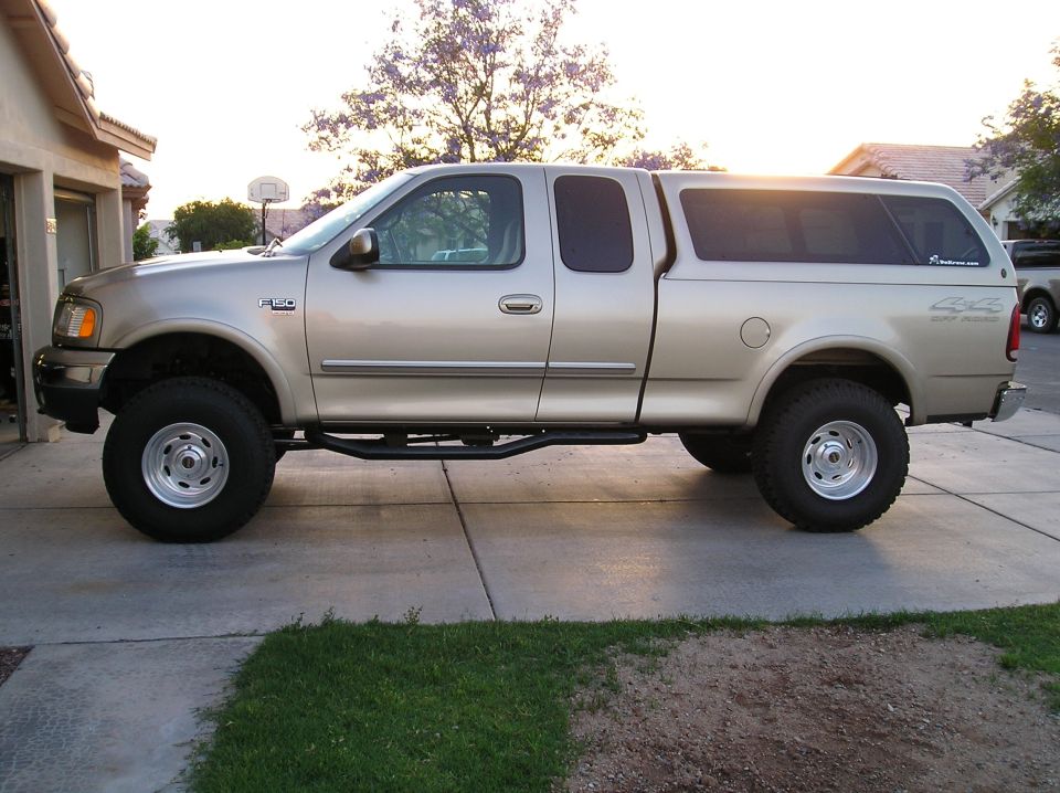 Tan lifted Ford F-150 extended-cab pickup with camper shell, side view in driveway.