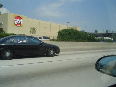 1993 Ford Taurus - A black 1993 Ford Taurus is seen driving on the highway, with several stickers visible on its rear window.