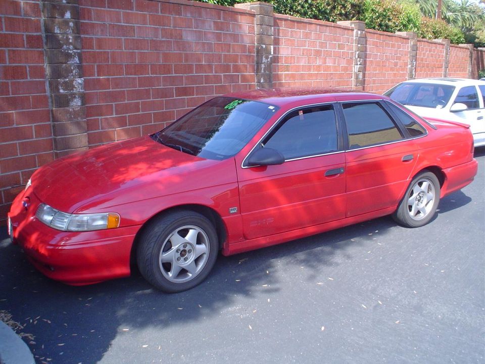 1993 Ford Taurus sedan parked on asphalt next to a brick wall. Another white car is visible behind it