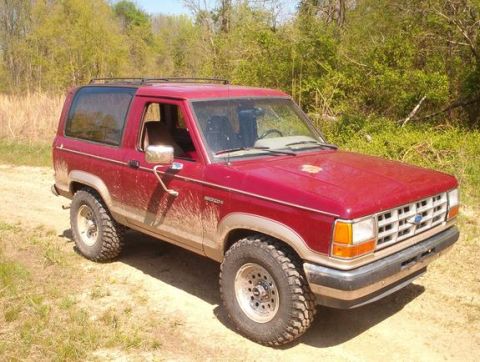 1989 Ford Bronco II - This red 1989 Ford Bronco II is shown in a three-quarter front view, parked on a dirt path with trees in the background.