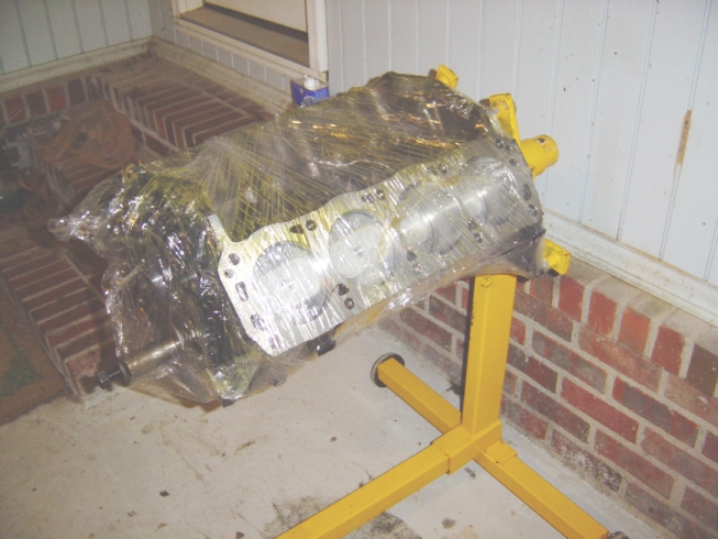 V8 short block on a yellow engine stand wrapped in plastic, bores visible