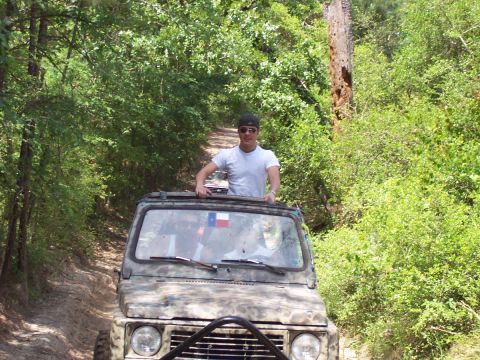 1989 Ford Bronco - A man stands up through the sunroof of a camouflage SUV as it navigates a wooded trail.