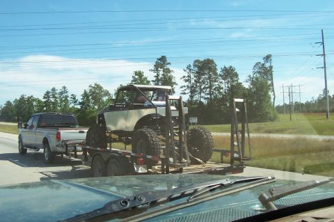 1989 Ford Bronco - A heavily modified off-road buggy with oversized tires is transported on a trailer behind a pickup truck on a highway.