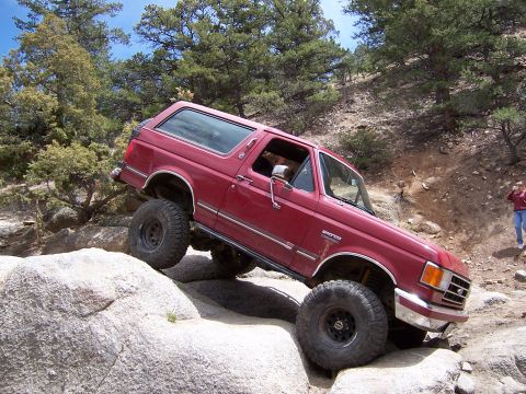 1991 Ford Bronco - A red 1991 Ford Bronco navigates a rocky off-road trail with large tires.