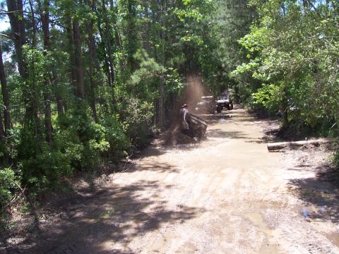 1989 Ford Bronco - Several off-road vehicles navigate a muddy trail surrounded by dense trees.