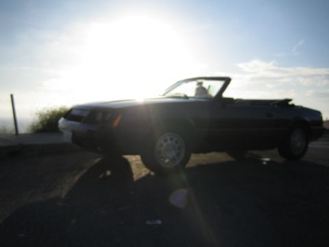 1985 Ford Mustang - A dark convertible car is seen from a three-quarter front angle against a bright sky.