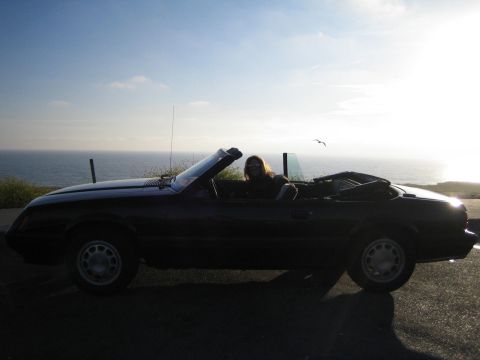 1985 Ford Mustang - A person enjoys a drive in a dark 1985 Ford Mustang convertible with the ocean in the background.