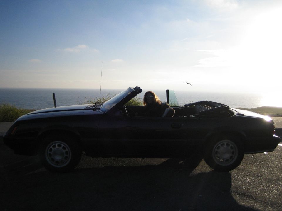 Dark 1985 Ford Mustang convertible side view with a person driving, ocean backdrop