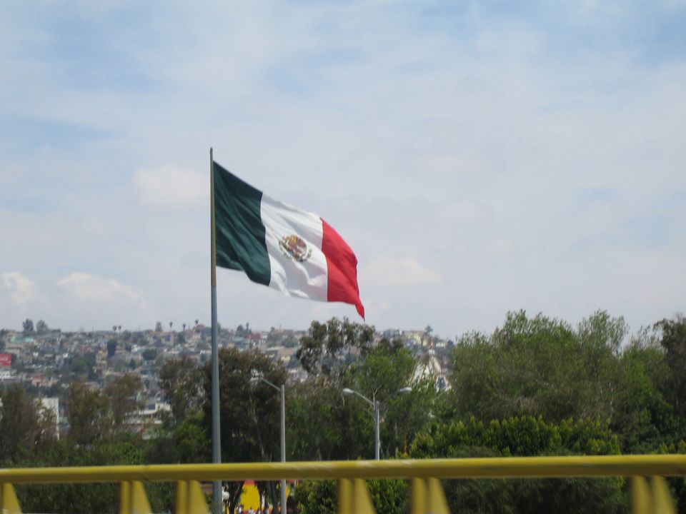 Large Mexican flag on tall pole above trees with hillside city in background