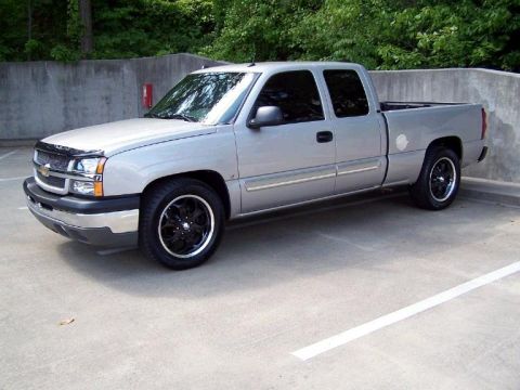 2004 Chevrolet Silverado 1500 - A silver pickup truck is parked in a lot with trees and a concrete wall in the background.