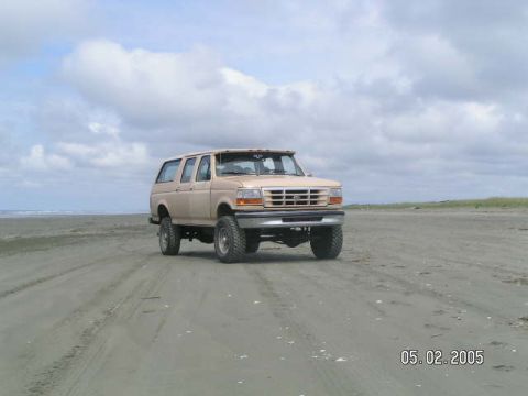 1988 Ford C-250 (Centurion Conversions) - A lifted tan Ford Bronco is parked on a sandy beach under a cloudy sky.