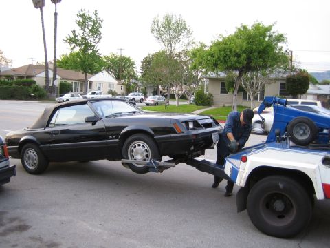 1985 Ford Mustang - A black 1985 Ford Mustang convertible is loaded onto a tow truck on a residential street.