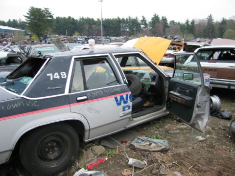 1991 Ford LTD Crown Victoria - A damaged 1991 Ford Crown Victoria LTD, marked with "749", sits in a junkyard with its door open.