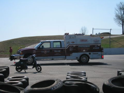 1991 Ford LTD Crown Victoria - A service truck with racing team decals is parked on a track next to a scooter, with tires in the foreground.