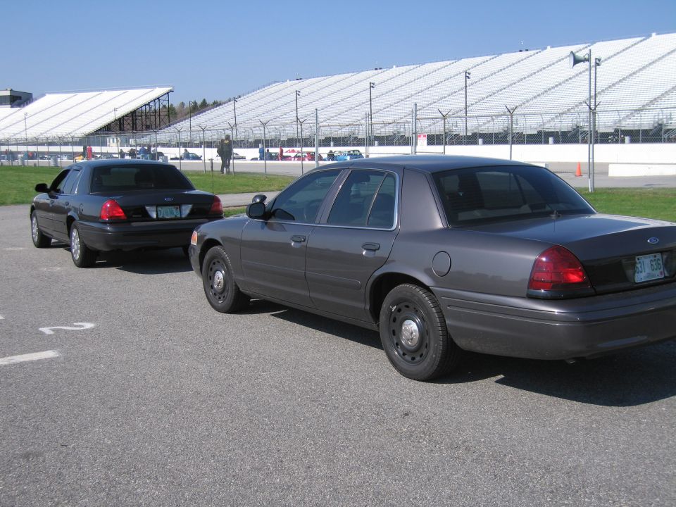 Two dark Ford Crown Victoria sedans parked on asphalt with grandstands in the background