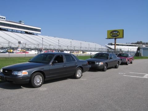 1991 Ford LTD Crown Victoria - Several sedans are parked in a line on asphalt, with a large grandstand and a NASCAR billboard visible.