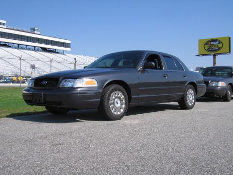 1991 Ford LTD Crown Victoria - A dark gray Ford Crown Victoria sedan is parked on asphalt with a racetrack grandstand in the background.