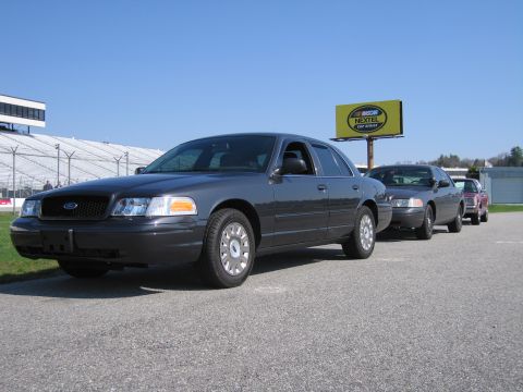 1991 Ford LTD Crown Victoria - Several dark sedans are parked in a line on a sunny day, with a NASCAR billboard visible.