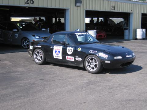 1991 Ford LTD Crown Victoria - A black Mazda Miata with racing decals is parked in front of garages, with a silver Ford Focus behind it.