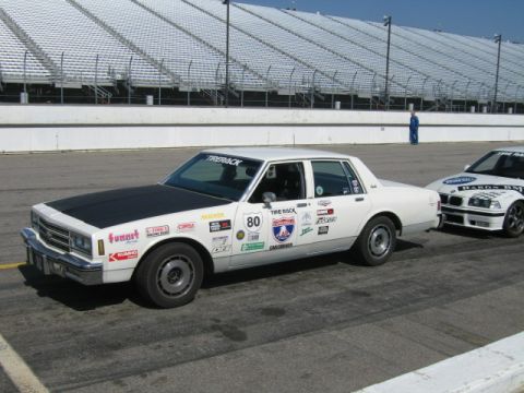 1991 Ford LTD Crown Victoria - White sedan with racing stickers and a black hood sits on a track next to another vehicle.