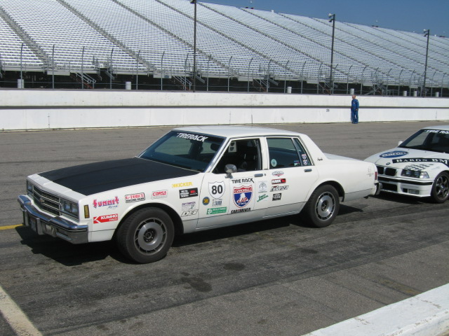 White sedan with racing stickers and black hood on a track, another car visible behind it