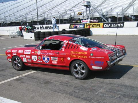 1991 Ford LTD Crown Victoria - A red 1965 Ford Mustang race car, adorned with many sponsor decals, sits on a track near grandstands.