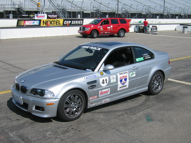 Silver BMW M3 coupe with racing decals and number 41 on a racetrack, red SUV in background