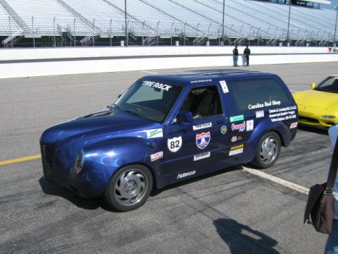 1991 Ford LTD Crown Victoria - A blue custom van with racing decals is parked on a track next to a yellow car, with grandstands in the background.