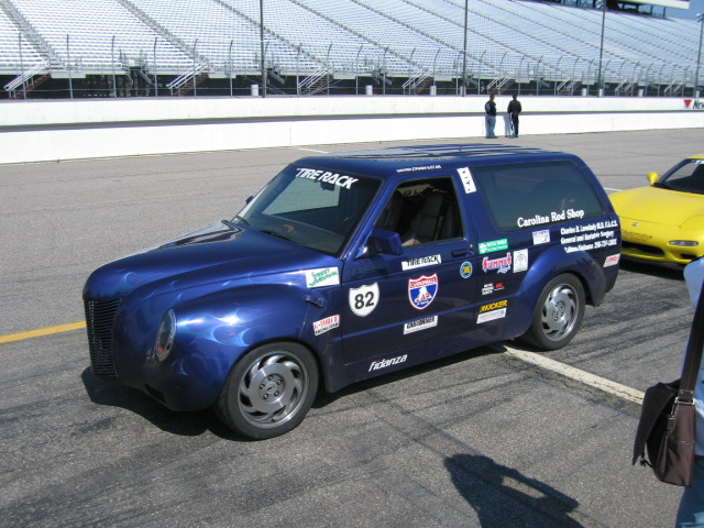 Blue custom van with flame decals and numerous racing stickers, seen from the front-left at a racetrack