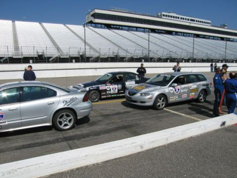 1991 Ford LTD Crown Victoria - Three cars are lined up on a racetrack, with spectators and grandstands visible in the background.