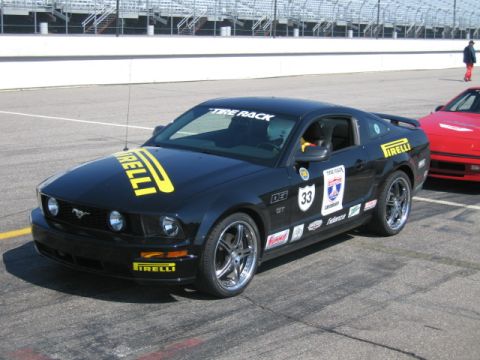 1991 Ford LTD Crown Victoria - Black 2005 Ford Mustang GT with racing decals parked on a track, with a red car in the background.
