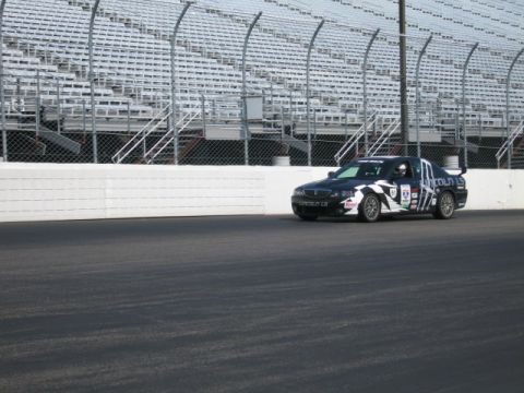 1991 Ford LTD Crown Victoria - A dark blue Lincoln LS race car with white checkered graphics is on a racetrack with grandstands in the background.