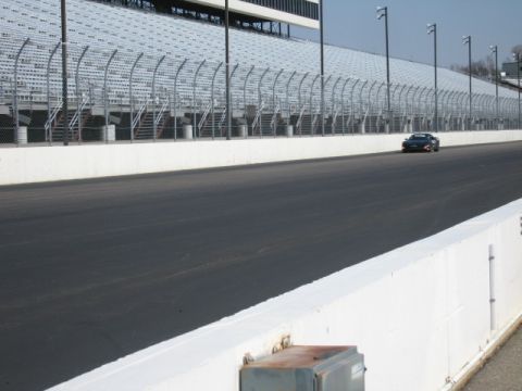 1991 Ford LTD Crown Victoria - A lone black sports car navigates a race track, with empty grandstands lining the perimeter.