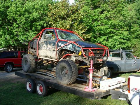 1985 Ford Bronco - A heavily modified truck with a roll cage is loaded onto a trailer, with other vehicles visible in the background.