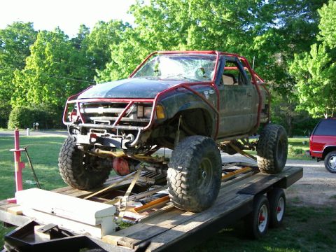 1985 Ford Bronco - This heavily modified off-road truck, with a custom roll cage and oversized tires, is loaded onto a trailer, with a red SUV visible in the…